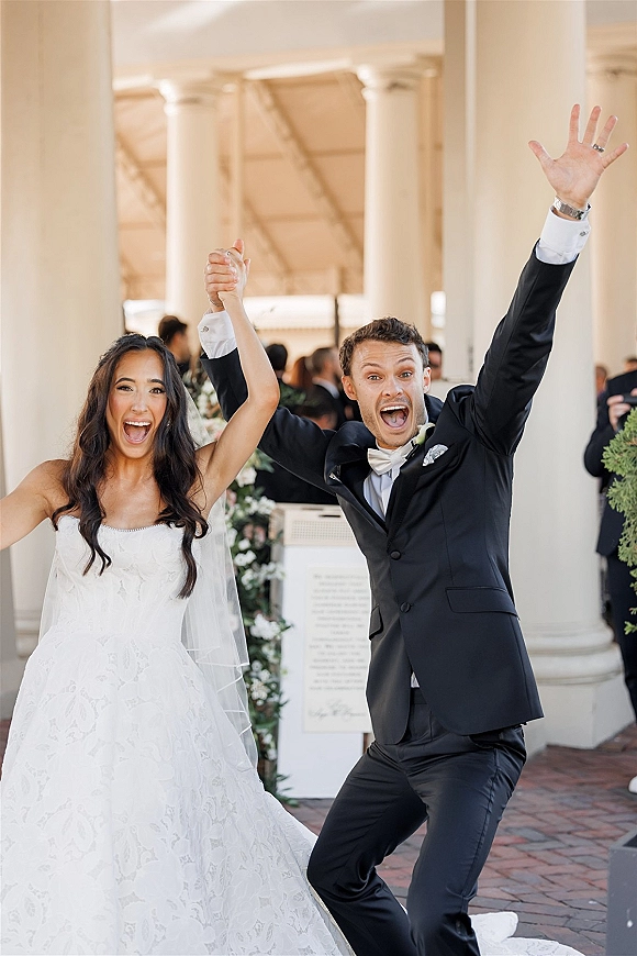 Recessional moment as bride and groom cheering with raised hands, just married, walking a brick walkway between white columns and guests