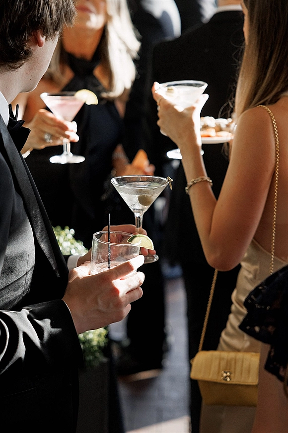 Wedding cocktail hour guests with cocktails holding martini glasses with lime wedge garnish, satin dress and tuxedo in soft indoor light