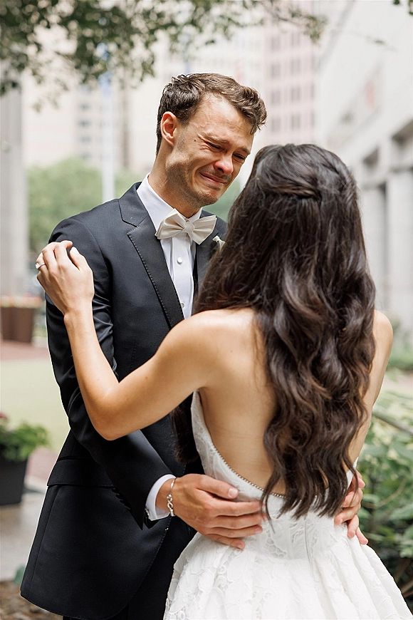 Wedding first look as a groom crying first look hugs the bride in a lace dress on a leafy city sidewalk among buildings and trees