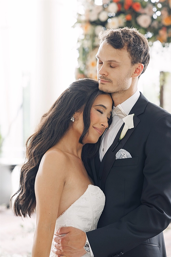 Couple portrait of bride and groom embrace, her strapless lace wedding dress against his black tuxedo in soft indoor light with blurred florals