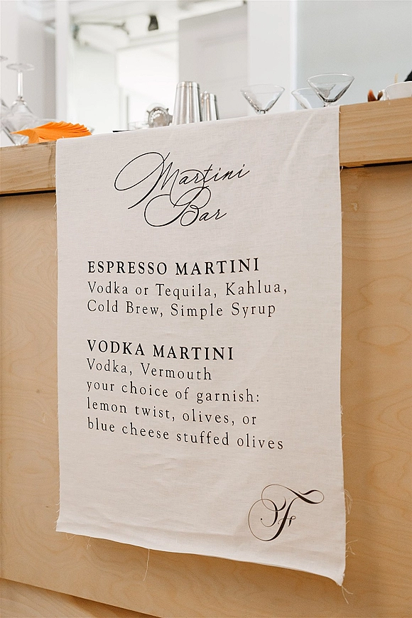 Wedding bar menu fabric sign with calligraphy lettering on a wooden bar, styled with martini glasses, shaker and napkins in a white interior