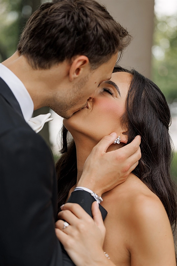 Wedding kiss portrait of bride and groom kiss close up as he cradles her face, tuxedo and bow tie against outdoor greenery and a column
