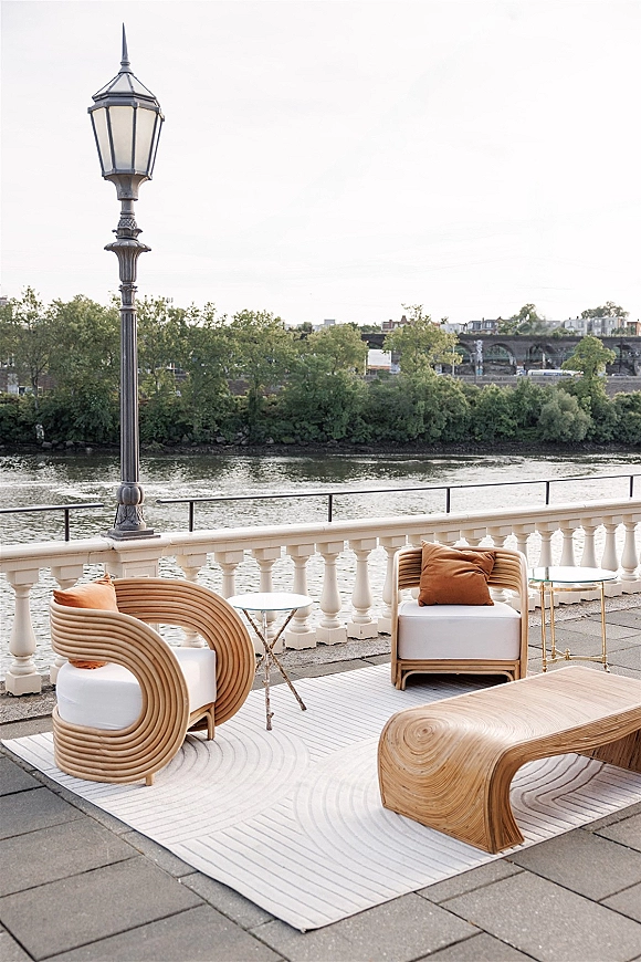 Lounge seating area with woven chairs and rust pillows around a curved wood coffee table on a riverfront terrace with bridge views