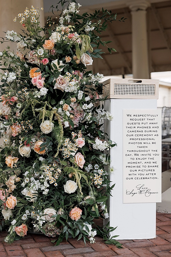 Wedding signage with an unplugged ceremony sign on a white pedestal, framed by rose and daisy florals and greenery on a covered patio