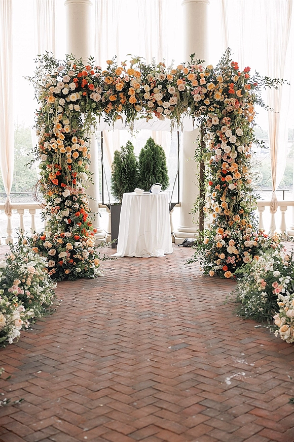 Wedding ceremony arch with roses and greenery framing white drapery, set before tall windows and white columns on a brick floor