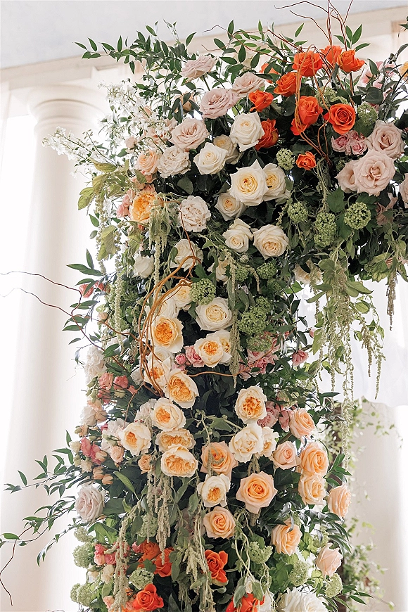 Wedding floral installation with cascading rose flowers and greenery on white columns, framed by tall indoor windows in soft natural light