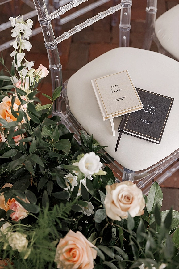 Wedding vow books with ribbon bookmarks resting on clear chiavari chairs, framed by roses and greenery above a brick floor