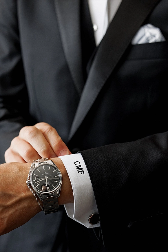 Groom accessories including a groom wristwatch on a French cuff shirt with cufflink, watch chain, and pocket square in an indoor setting