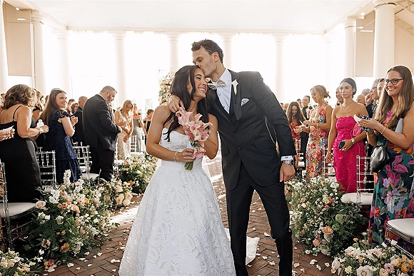 Recessional moment as bride and groom walk the brick aisle, groom kissing her forehead as she holds calla lilies, guests stand by columns.