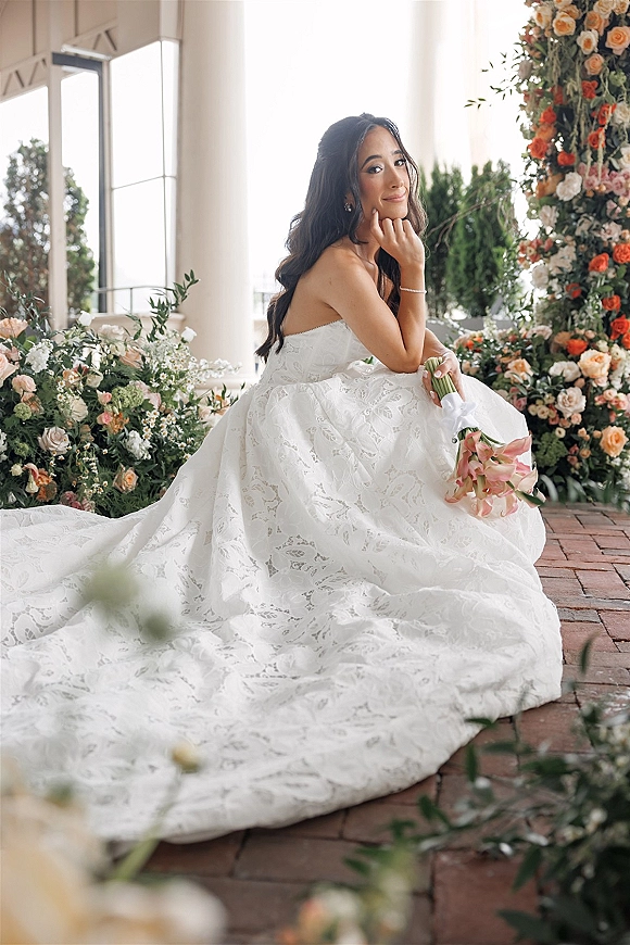 Bridal portrait of a bride sitting in a strapless lace wedding dress, holding a pink calla lily bouquet beneath a floral arch in a brick courtyard