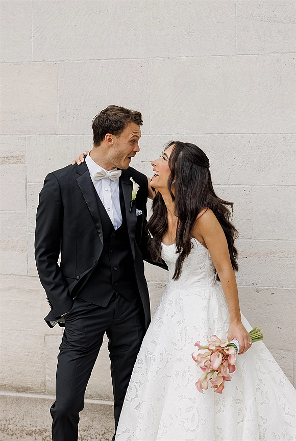 Couple portrait of bride and groom laughing, her bouquet in hand, in a strapless lace wedding dress and black tuxedo by a stone wall