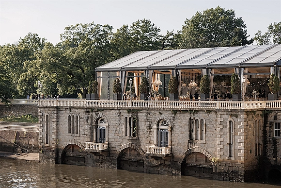 Wedding reception tent with clear top tent, chandeliers, and string lights over banquet tables on a stone terrace by the river