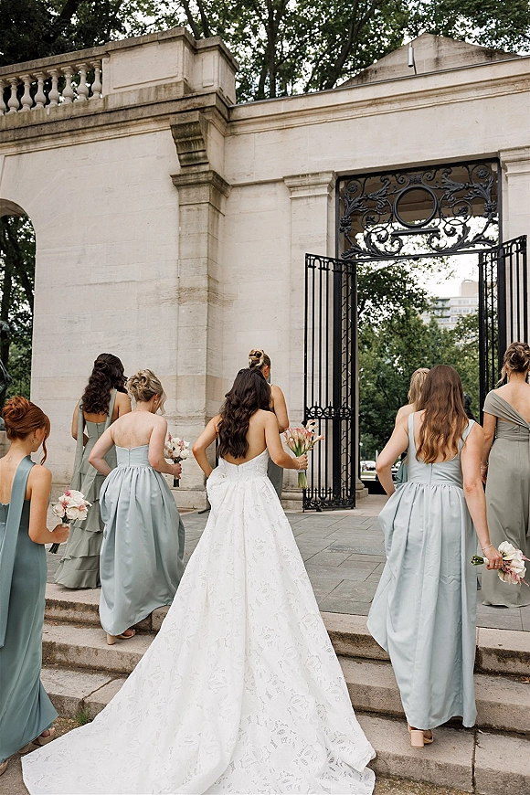 Bridal party with bride with bridesmaids walking up stone steps, her long train trailing as they hold bouquets by an iron gate under trees