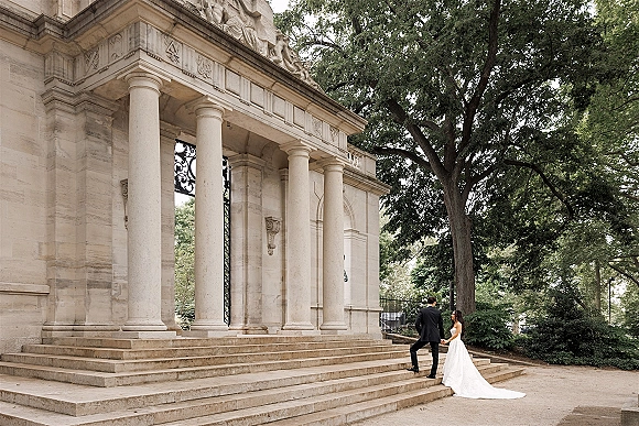 Couple portrait of bride and groom holding hands as he leads her up steps, her strapless gown train flowing by stone columns