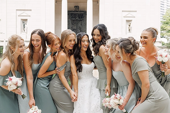 Bridesmaid group photo with bride with bridesmaids laughing, holding white and blush bouquets outside a stone building with columns