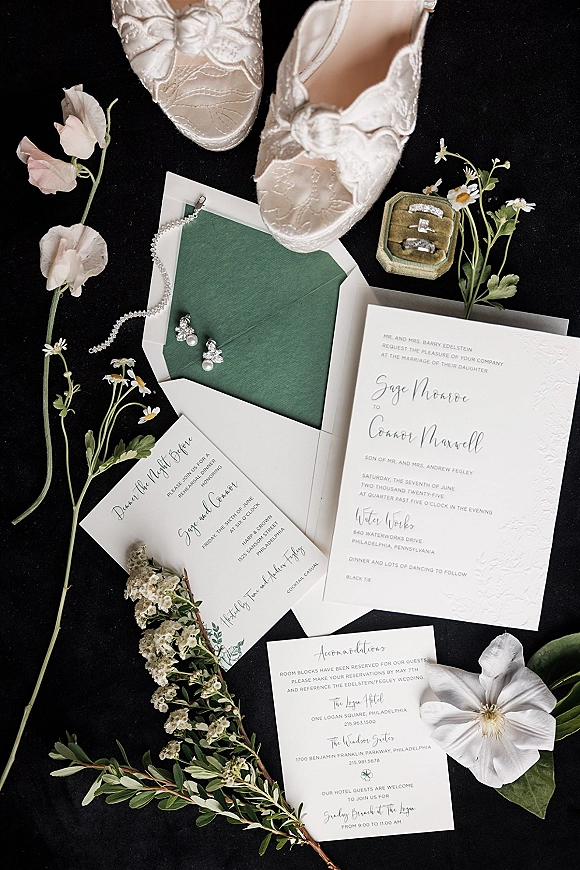 Invitation suite wedding invitation flat lay with green envelope liner, rings in velvet box, bridal shoes and pearls on black fabric backdrop
