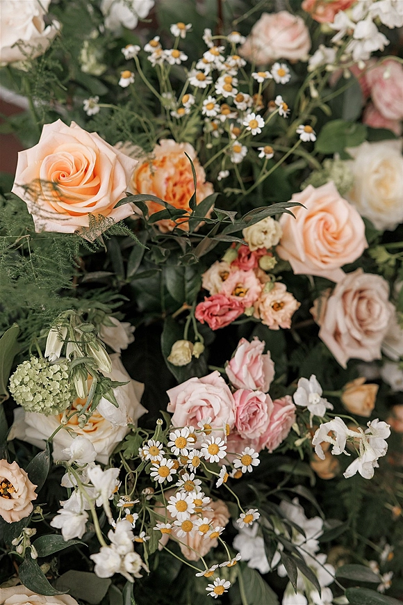 Wedding florals with a peach rose arrangement of blush and pink roses, white daisies, and greenery against a soft blurred floral backdrop