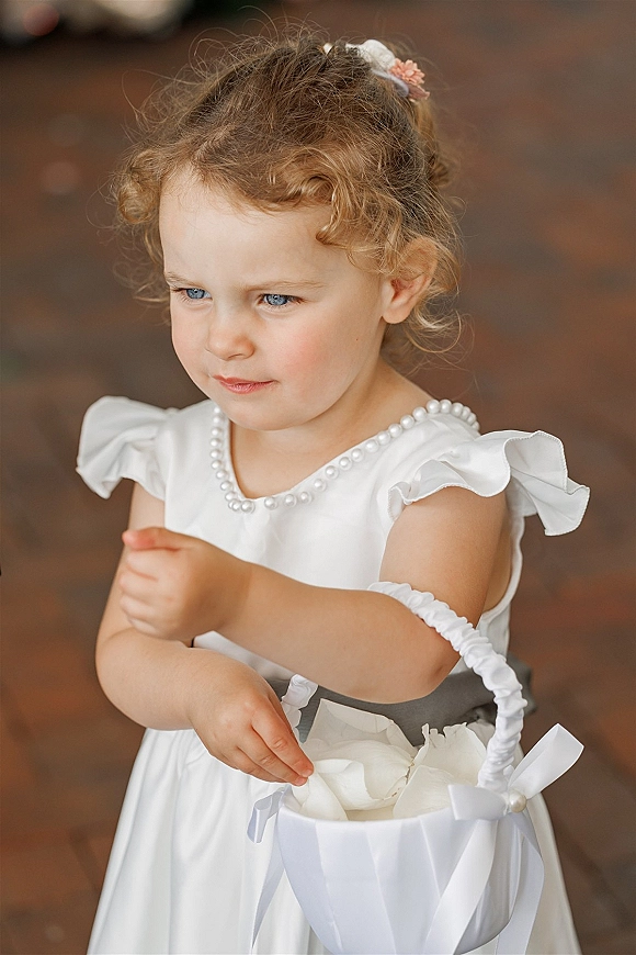Flower girl portrait of a toddler holding a petal basket, wearing a white dress with pearls and a flower clip on a blurred indoor floor