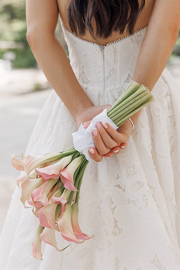 Bridal bouquet of blush calla lilies held behind the bride’s back, wrapped in white ribbon with lace dress detail on an outdoor walkway
