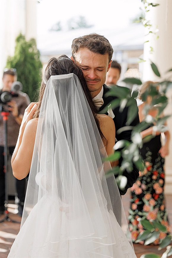 First look moment as bride in a wedding dress with veil hugs groom in tuxedo on an outdoor patio with white columns and guests nearby