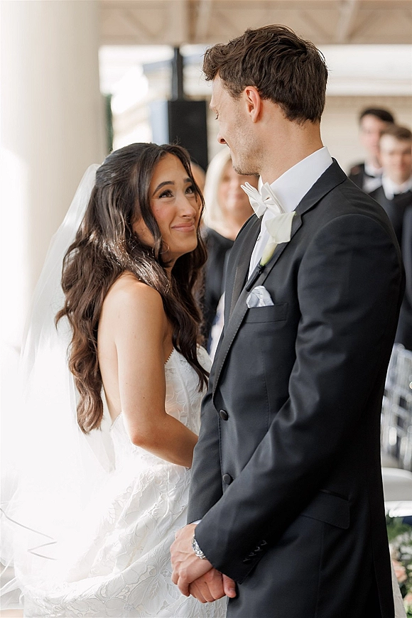 Ceremony moment as bride looks at groom while holding hands, her long veil draped over lace dress in soft indoor light with guests behind