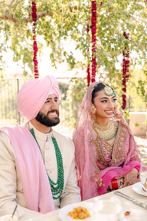 Couple portrait of an Indian wedding couple seated at an outdoor garden lunch table, bride in embroidered lehenga and groom in pink turban under floral garlands