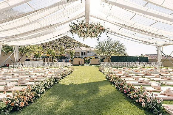 Ceremony setup with outdoor wedding ceremony seating under a clear top tent, floral chandelier, draped canopy, string lights, and mountain lawn backdrop