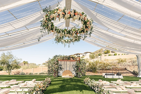 Ceremony setup for an outdoor wedding ceremony with draped canopy, hanging floral chandelier, and floor cushions on a grass lawn under blue sky