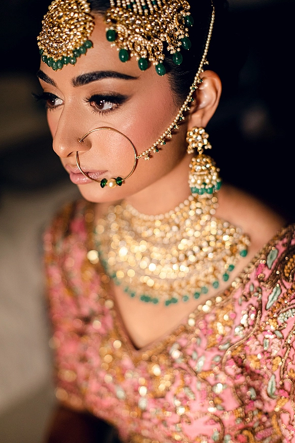 Bridal portrait of a South Asian bride in gold headpiece and nath nose ring with chain, green gemstone necklace, against a dark background