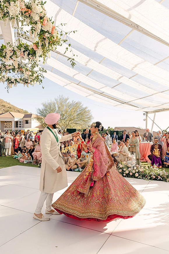 Wedding first dance as bride twirls in a pink lehenga with bangles beside groom in cream sherwani and turban under a floral canopy on lawn