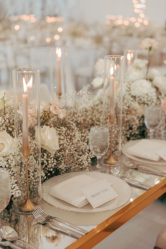 Reception tablescape with wedding table centerpiece of white roses and baby’s breath garland, gold taper candles on mirrored tabletop under string lights