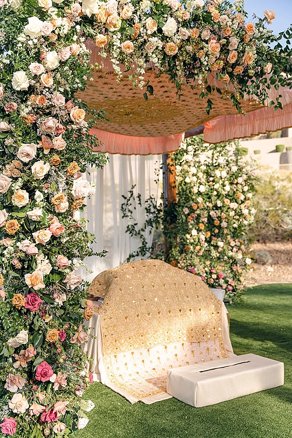 Wedding lounge setup with outdoor wedding lounge draped canopy, rose and greenery floral arch, and floor cushions on a garden lawn under blue sky