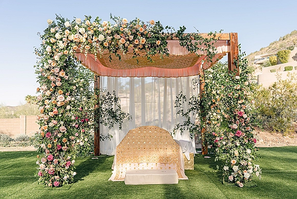 Wedding ceremony backdrop with a floral arch backdrop of roses and greenery, sheer drapes and gold canopy fabric on a lawn by desert hills