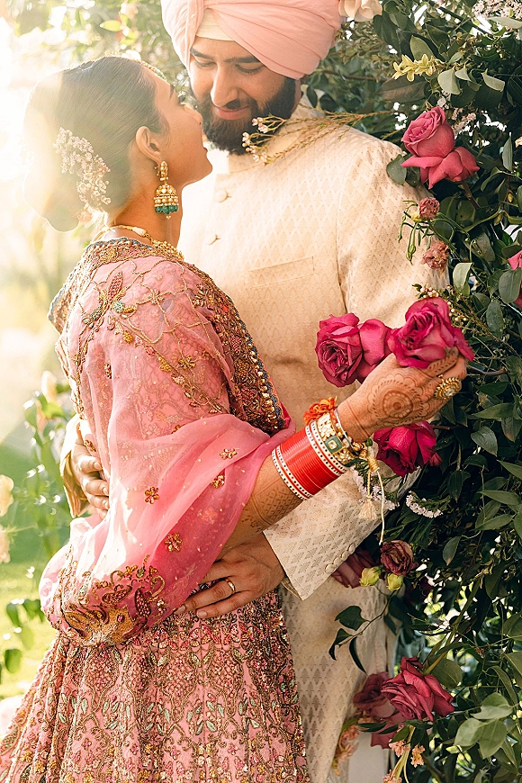 Couple portrait of a sikh wedding couple embracing under a pink rose floral arch, bride shows mehndi and red bangles in sunlight