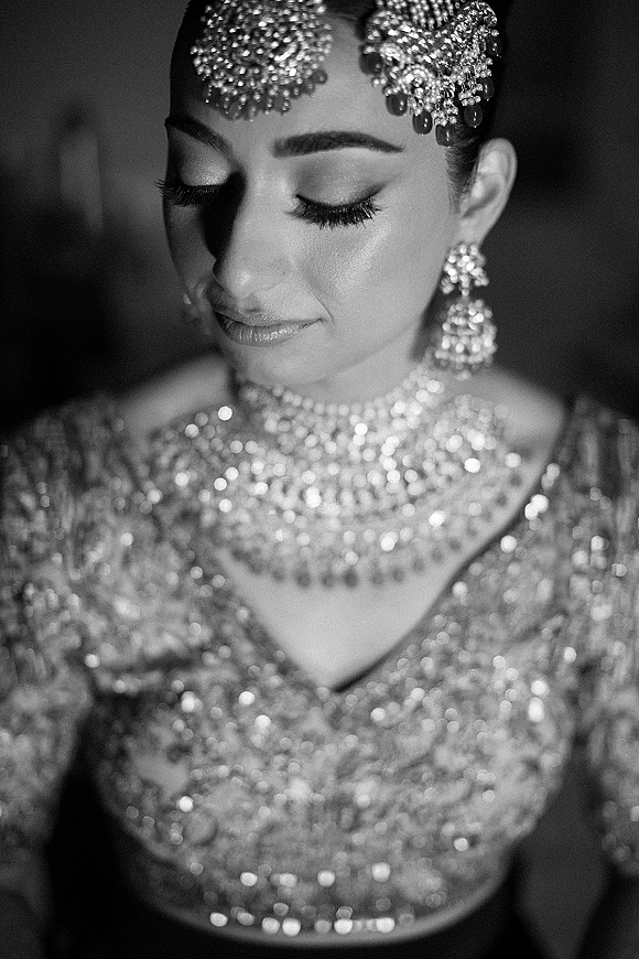 Bridal portrait in black and white showing crystal headpiece, chandelier earrings, and statement necklace against a dark indoor background