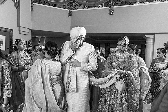 Wedding ceremony moment as groom in sherwani and turban stands with bride in embroidered lehenga and jewelry among indoor guests and columns