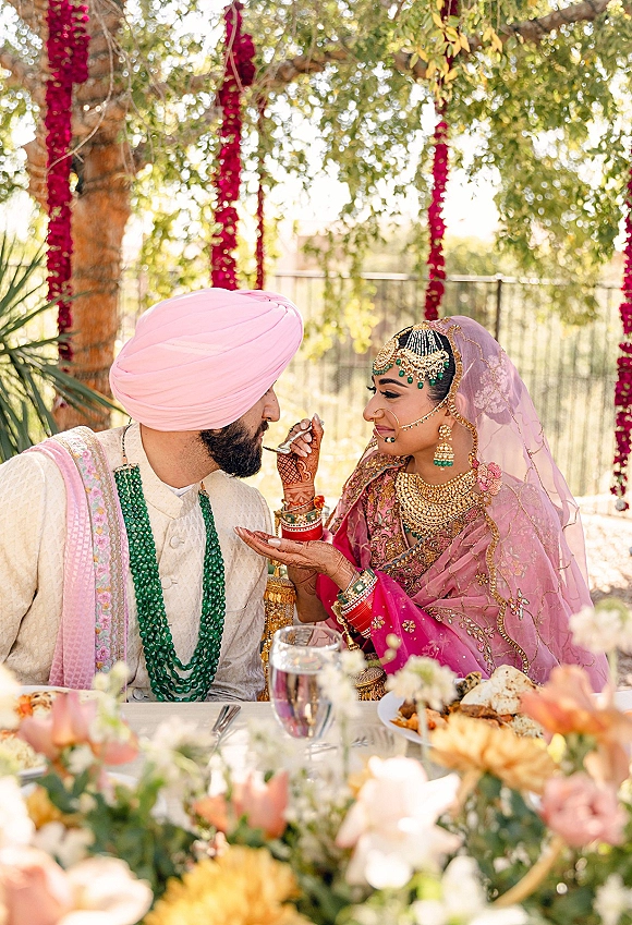 Ceremony ritual moment as a Sikh wedding couple shares a feeding tradition at a table outdoors under sunlight and hanging garlands