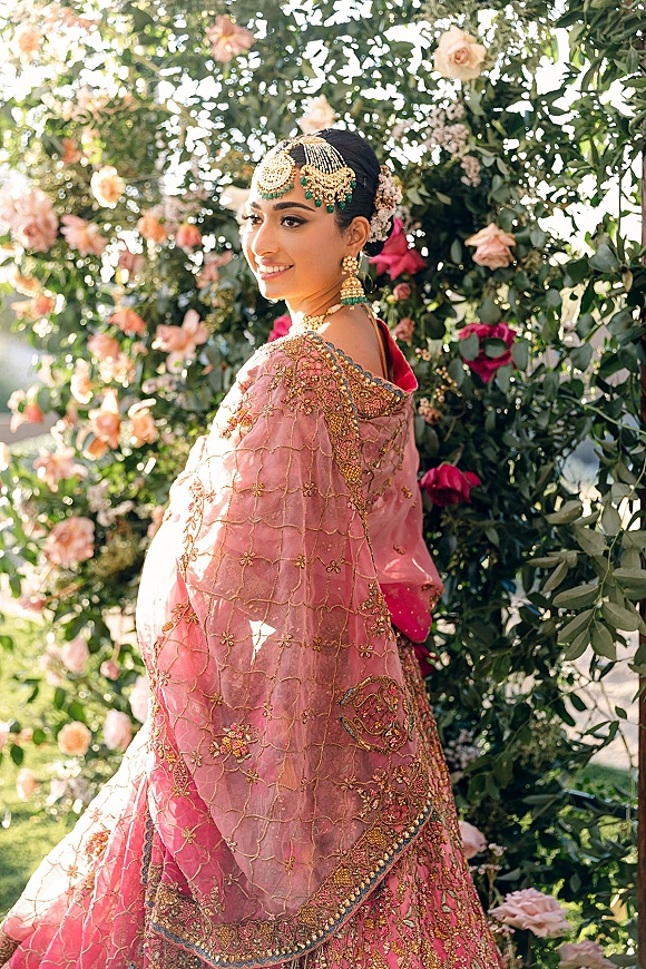 Bridal portrait of a south asian bride in a pink lehenga with gold embroidered dupatta, maang tikka and jhumkas, posed by a rose flower wall outdoors