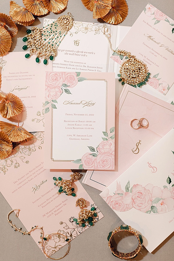 Wedding invitation suite with floral wedding invitations, blush envelopes, monogram emblem, gold foil edging, rings and emerald jewelry on neutral tabletop