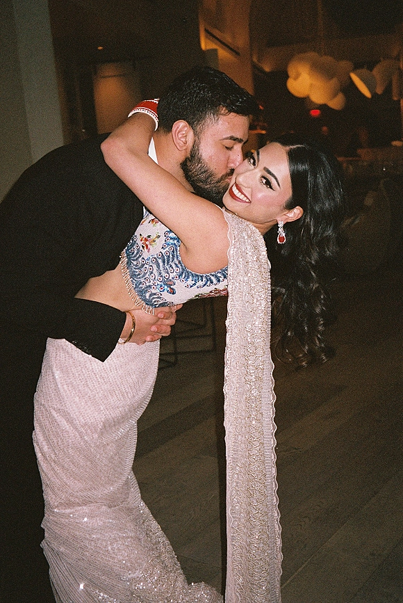 Wedding kiss portrait of groom dipping his bride in a bridal lehenga, embroidered dupatta and red lipstick in a dim indoor reception space
