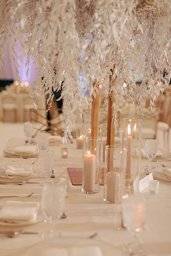 Reception tablescape with wedding table centerpiece of white florals on a wooden stand, candlelit with glass hurricanes and taper candles under hanging greenery
