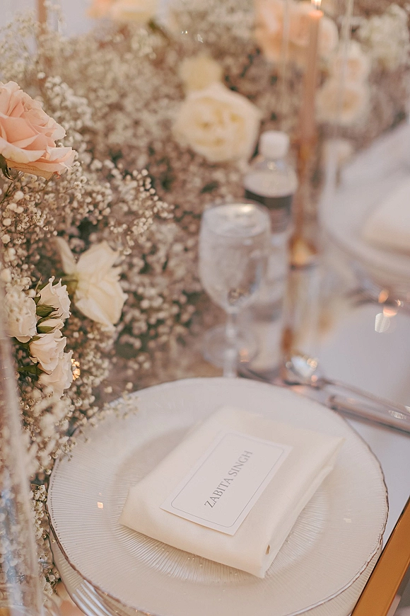 Reception tablescape with a wedding place setting featuring a white plate, folded napkin and place card, glass goblet, blush roses, and taper candle