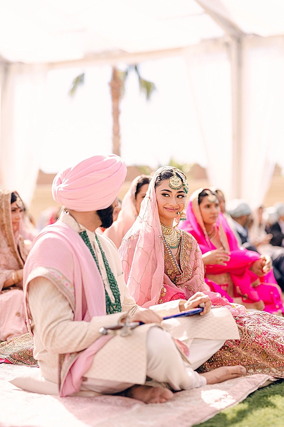Ceremony moment at a Sikh wedding ceremony with bride in pink lehenga and groom in turban seated on rugs under a tent canopy, guests behind