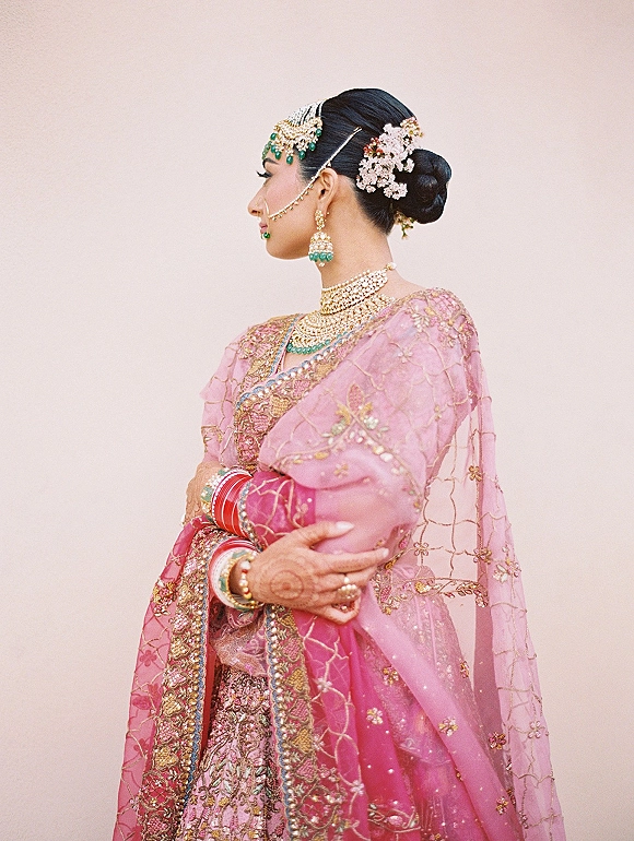Bridal portrait of an Indian bride in a pink embroidered lehenga with gold jewelry, gajra bun, and mehndi hands on a neutral studio backdrop
