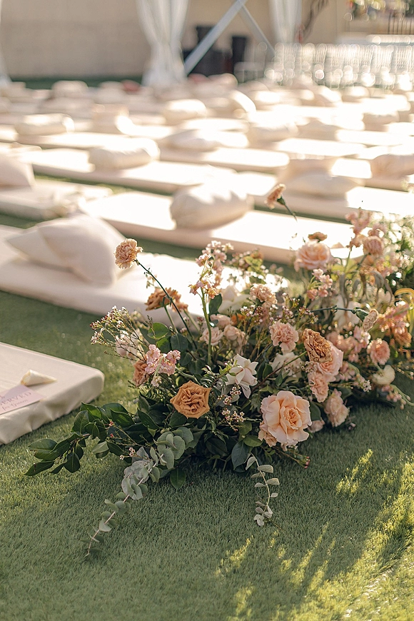 Ceremony aisle decor with a ground floral aisle of blush and peach roses, eucalyptus greenery, floor cushions and place cards on a grass lawn