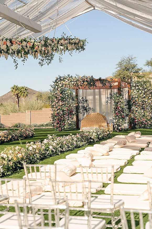 Ceremony setup with a floral arch and draped canopy over a white aisle runner, clear chairs and cushions on a lawn with palms and desert mountains