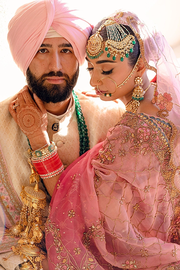 Couple portrait of a sikh wedding couple outdoors under bright sky, bride in pink dupatta veil and gold jewelry beside groom in turban