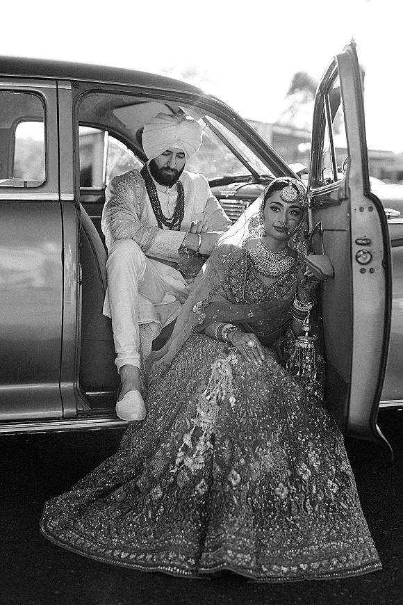 Couple portrait of bride and groom in car, bride in embroidered lehenga as groom in turban sits by open door on street