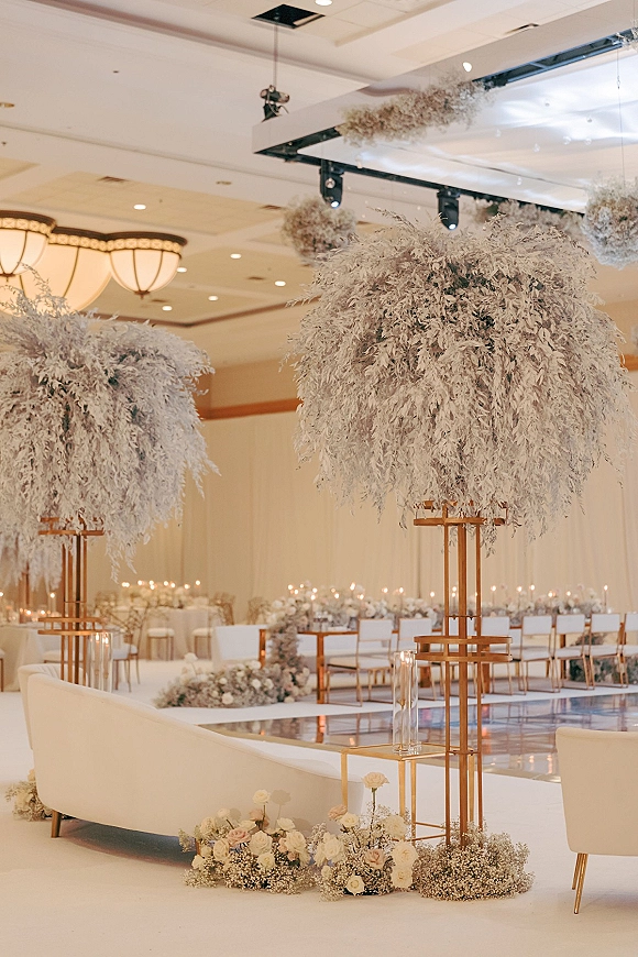 Ceremony setup with white aisle runner lined by floral arrangements and glass cylinder candles, with hanging florals in a ballroom