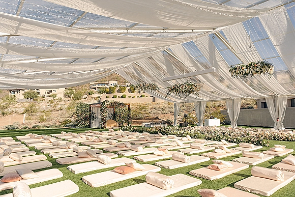 Wedding ceremony setup with outdoor ceremony canopy, sheer draping and string lights over lounge cushions on a desert lawn with hills backdrop